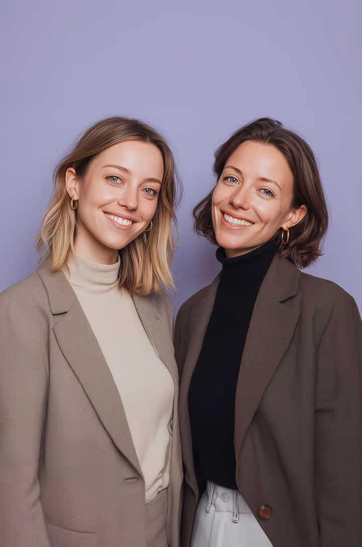 Two smiling women wearing blazers, one in beige with a light turtleneck and the other in brown with a black turtleneck, standing against a purple background.