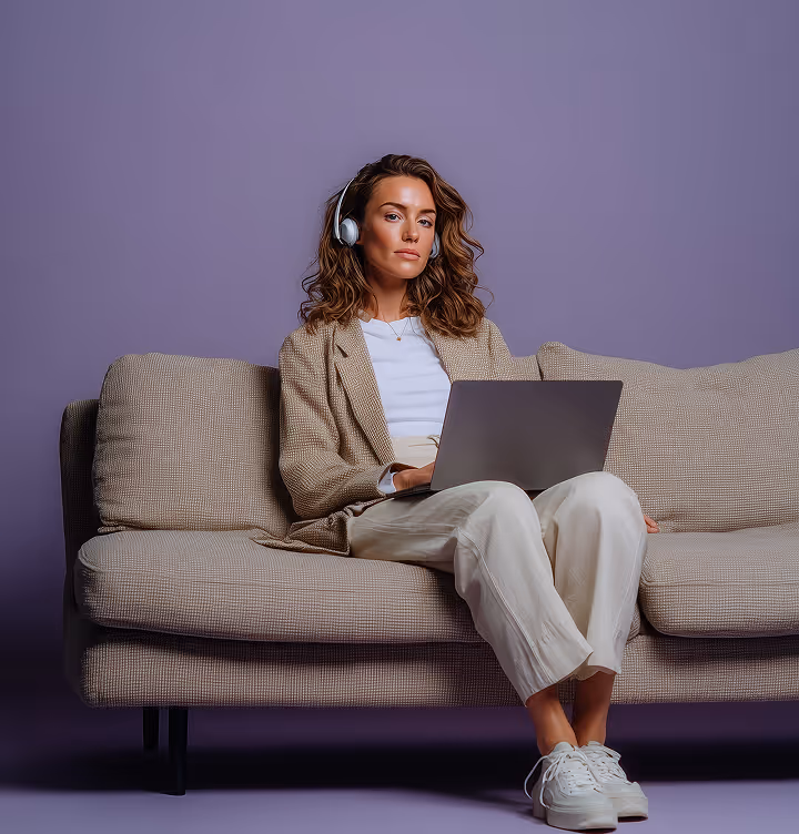 Woman with curly hair wearing headphones, sitting on a beige sofa and working on a laptop against a purple background.